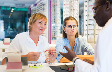Young woman with elderly mother looking for glasses in optical shop, asking for advice from qualified African-American optometrist