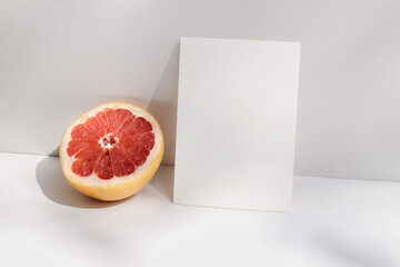 Modern summer stationery still life scene. Closeup of cut grapefruit fruit on white table background in sunlight. Blank paper card, invitation mockup scene lean on beige wall, long shadows.