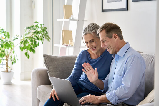 Happy Old Senior Grandparents Couple Waving Hand Making Online Video Call Enjoying Family Virtual Meeting Digital Webcam Videocall Chat Talking, Looking At Laptop Computer Sitting On Couch At Home.