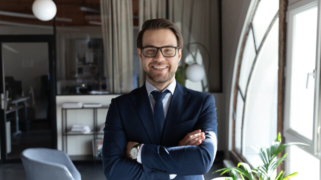 Head shot smiling confident young businessman executive wearing glasses and suit standing in modern office room, happy successful entrepreneur employee intern with arms crossed looking at camera