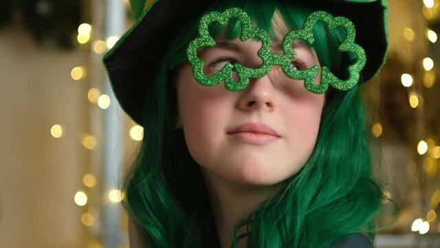 Saint Patrick's day celebration concept. Beautiful girl with green hair wearing green hat and glasses in the shape of a clover. Closeup portrait.