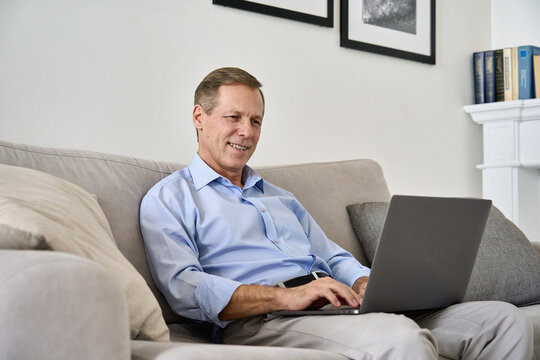 Smiling Happy Middle Aged 50s Old Senior Man Using Laptop Computer Technology Working Online, Surfing Internet, Typing Email, Shopping In Ecommerce Store Relaxing With Device Sitting On Couch At Home.