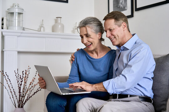 Happy old senior retired grandparents using laptop sitting on couch at home. Smiling mature 50s family couple holding computer technology looking at device e shopping or banking online together. - Powered by Adobe