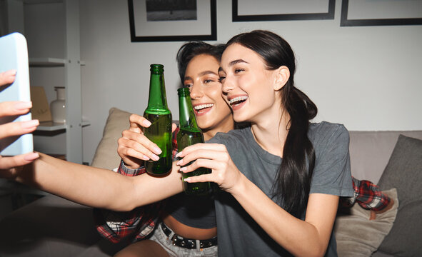 Two happy young girls drinking beer having virtual party remote meeting chat looking at smartphone video calling social distance friends online celebrating together having fun late at night at home. - Powered by Adobe