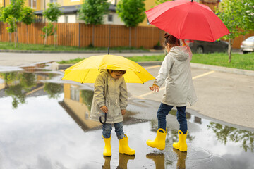 Two happy sisters in yellow rubber rain boots play with bright umbrellas in the street after the rain