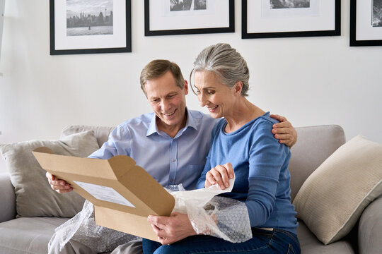 Happy older mature couple customers unpacking parcel sitting at home on couch. Senior middle aged grandparents opening online store order receiving gift in postal delivery shipping box.