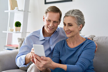 Middle aged 50s senior couple holding device looking at smartphone using mobile apps tech, shopping in ecommerce application, ordering delivery, buying goods online on cell phone sit on sofa at home.