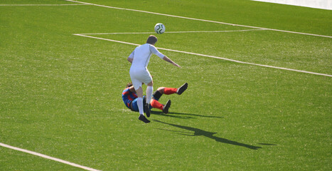 Dois jogadores a disputar a posse de bola um caído no chão, o outro a saltar para não pisar o que está no chão enquanto ao mesmo tempo tenta cabecear a bola na direcção da baliza