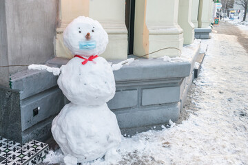 A snowman in a medical mask stands on an empty sidewalk. City street during winter lockdown. Winter...