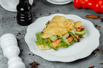 Croissant sandwich with red fish, avocado, fresh vegetables and arugula on black shale board over black stone background. Healthy food concept