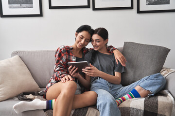 Happy young lesbian couple hugging having fun using digital tablet relaxing on couch at home. Two...