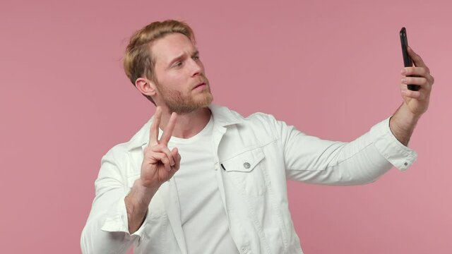 Funny bearded young man 20s in white clothes isolated on pink background studio. People lifestyle concept. Doing selfie shot on mobile phone showing biceps muscles thumb up victory sign point himself