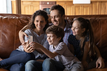Smiling young Caucasian family with two small children have fun talk on video call on smartphone. Happy parents relax at home with little kids watch funny video on cellphone. Technology concept.
