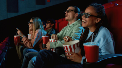 Happy young woman wearing glasses, eating popcorn while watching movie in cinema auditorium...