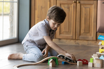Small 6s Caucasian boy sit on warm floor at home play with cars and wooden road. Happy little child have fun feel playful with toys, involved in game activity. Development, entertainment concept.