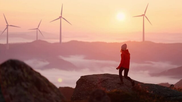 Young woman in orange jacket running up on top of mountain summit at sunset, raises arms into air, happy and drunk on life and happiness. Girl stand against landscape with wind turbines on background