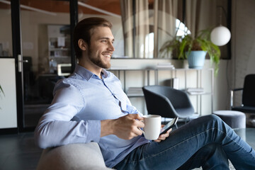 Smiling businessman holding cup, sitting in armchair in modern office room, enjoying break, looking to aside, successful entrepreneur visualizing good future, dreaming about new opportunities