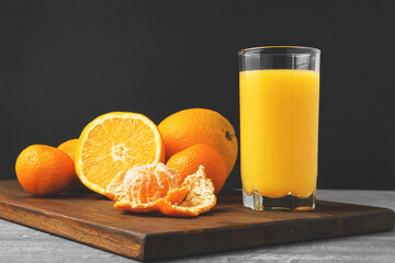 Glass of fresh orange juice, whole and sliced fruits on wooden board on dark background with copy-space