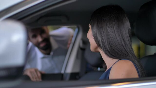 Close-up of happy brunette female testing buying car from inside during visit auto dealership. Excited woman sitting in driver's seat while her husband looking in car window blurred on background