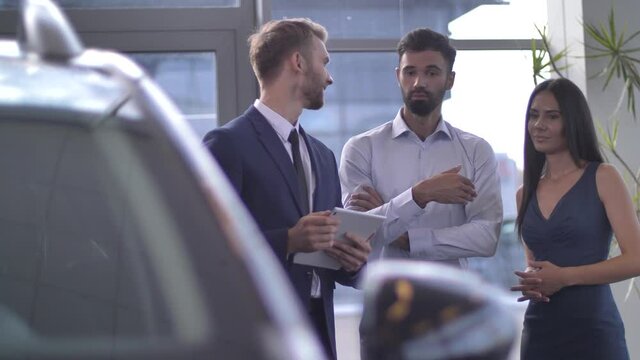 Confident salesman consulting couple of picky buyers visiting car showroom. Demanding customers using help of professional car dealer while choosing luxury automobile in dealership
