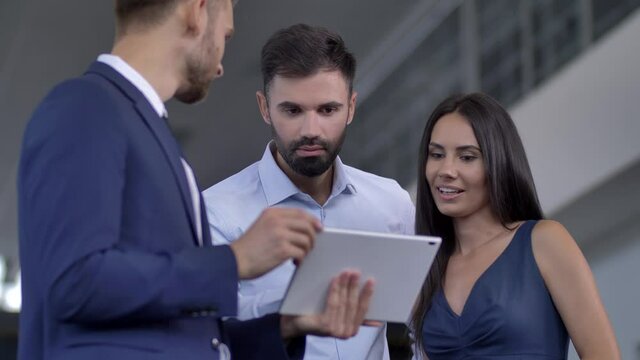 Close-up of professional salesman using tablet while showing couple of customers specifications of chosen car. Interested woman and man communicating with auto dealer and making choice at car showroom