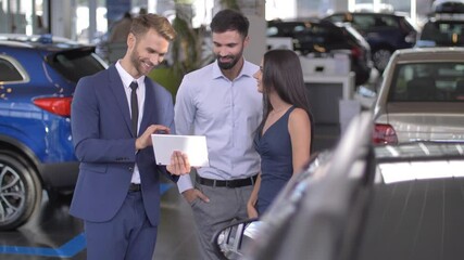 Smiling young couple discussing automobile specifications with salesmanwhile visiting car dealership. Happy husband and wife using consultation of auto dealer with tablet while choosing car - Powered by Adobe