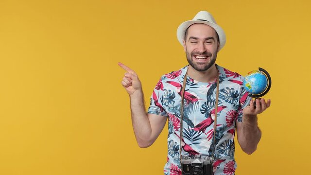 Smiling traveler tourist young man in hat isolated on yellow background. Passenger traveling on weekends. Air flight journey concept. Hold Earth world globe pointing finger aside doing winner gesture