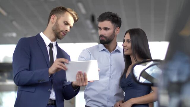 Serious handsome salesman with tablet consulting young couple visiting car showroom to buy new automobile. Professional auto dealer showing interested clients specifications of chosen car on tablet pc