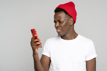 Confused African millennial hipster man looking at smartphone, annoyed by spam, intrusive adware, unpleasant message isolated on grey studio background. Black male has problem with cellphone, bad news