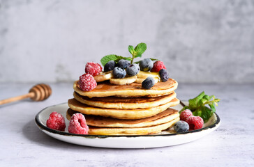 Homemade pancakes with banana, raspberries, blueberries and honey on a light kitchen background.