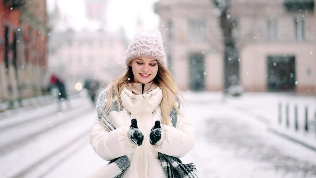 As the snow falls from the sky, a beautiful blonde woman starts playing, breathing clean air and dancing in the city street. Close up portrait of female in snowy holiday city. Snowing outdoors