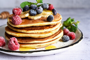 Homemade pancakes with banana, raspberries, blueberries and honey on a light kitchen background.