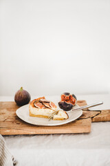 Slice of homemade fig cheesecake with fresh figs and fork on white plate over rustic wooden board, white wall at background, copy space. Healthy dessert, artisan sweet food concept
