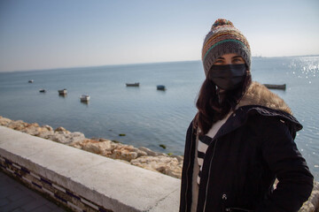 Beautiful girl with mask and hat next to the beach in winter with boats in the water
