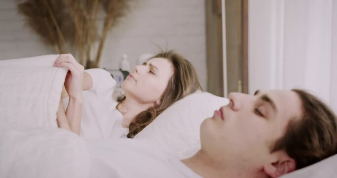 Family couple lying together in bed. Husband snoring loudly, preventing wife from falling asleep. Irritated woman staying awake, covering ears with pillow. Close-up shot.