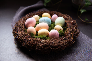 A wicker nest with moss and colored Easter eggs on gray linen fabric napkin and few green leaves on dark background. Dark mood style. Happy Easter holiday, selective focus