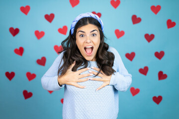 Young beautiful woman over blue background with red hearts looking happy, surprised, proud and excited, pointing to self