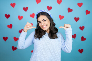 Young beautiful woman over blue background with red hearts looking confident with smile on face, pointing oneself with fingers proud and happy.