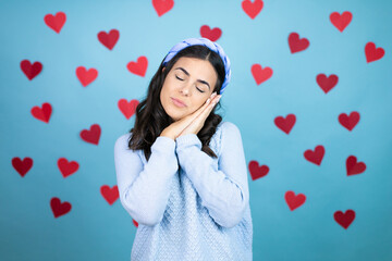Young beautiful woman over blue background with red hearts sleeping tired dreaming and posing with hands together while smiling with closed eyes.