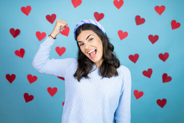 Young beautiful woman over blue background with red hearts showing arms muscles smiling proud