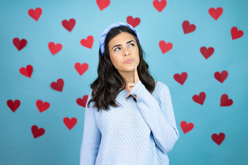 Young beautiful woman over blue background with red hearts with hand on chin thinking about question, pensive expression. smiling and thoughtful face. doubt concept.