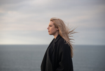 mujer rubia con abrigo negro mirando hacia el mar y tomando el aire en un día de invierno y cielo nublado