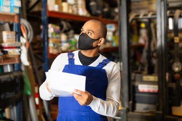 Warehouse male worker in protective mask and overalls counts the quantity of goods and writes in a notebook