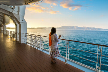A woman sips a drink on the deck of a cruise ship as the sun sets and the ship passes islands on...