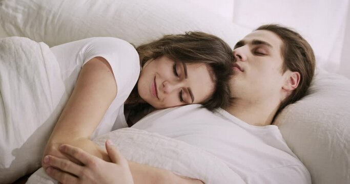 Young Caucasian couple enjoying good quality sleep, resting together in bed. Loving woman wife sweetly cuddling her husband, smiling.