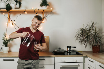 portrait of a happy European in the kitchen. A man pours water from a jug into a glass.