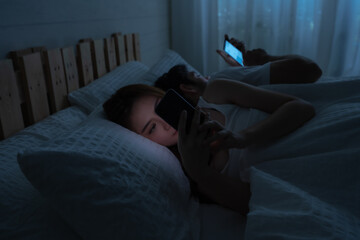 Asian woman and Caucasian man lay on bed use smartphone for social media and online at night. Copy space concept of diversity couple, relationship and communication problem, social media addiction.