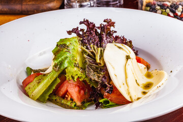 Italian caprese salad with sliced tomatoes, mozzarella cheese, pesto sauce, lettuce on white plate.