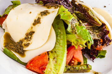 Italian caprese salad with sliced tomatoes, mozzarella cheese, pesto sauce, lettuce on white plate.
