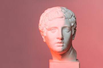 Frontal view of a white marble head of young man over red-lit pink background. On a square pedestal.